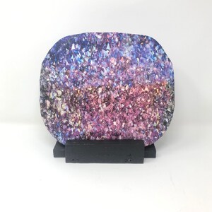 以下が含まれることがあります： 黒いスタンドの上に置かれた、紫と青のクリスタルジオード。ジオードは、虹色に輝き、光の中でキラキラと輝く、たくさんの小さなクリスタルでできています。