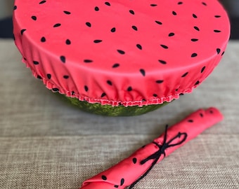 May include: A pink reusable food wrap with a watermelon print. The wrap is made of fabric and has a black elastic band around the edge. The wrap is rolled up and tied with a black string.