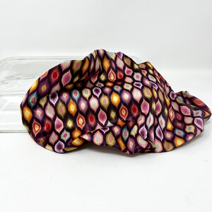 May include: A colourful fabric with a geometric pattern of teardrop shapes in shades of pink, orange, yellow, green, and blue on a dark purple background.