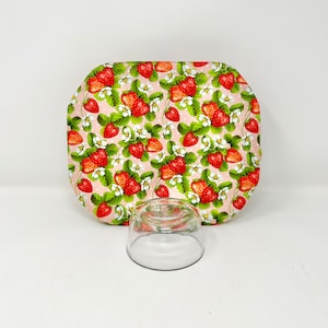 May include: A pink and white oval-shaped cake plate with a strawberry pattern. The plate is sitting on a white surface with a clear glass cake dome next to it.