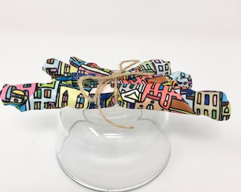 May include: A clear glass bowl with a colorful fabric tied around the top with twine. The fabric has a pattern of colorful buildings and lines.
