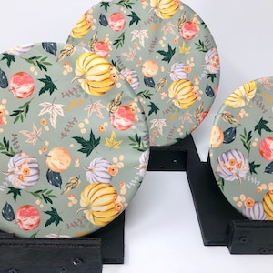 May include: Three round, green fabric placemats with a fall-themed print of pumpkins, leaves, and berries. The placemats are displayed on black wooden stands.