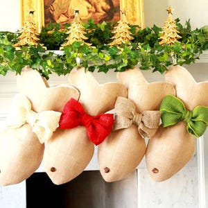 May include: Four burlap stockings with different coloured bows hanging on a white mantelpiece. The stockings are shaped like reindeer heads and have a brown burlap fabric with a white trim. The bows are red, green, brown, and white.