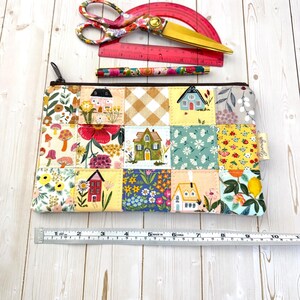 May include: A rectangular fabric zipper pouch featuring a patchwork design of various floral, house, and gingham patterns in yellow, blue, red, and green. A metal ruler below shows the pouch is approximately 10 inches wide. Includes scissors and a pen.