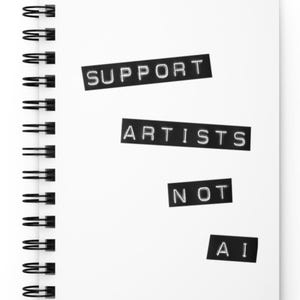 Puede incluir: Un cuaderno blanco con espiral con las palabras "SUPPORT ARTISTS NOT AI" en letras negras. El cuaderno está abierto y el texto está dispuesto en la página en una fuente clara y en negrita.