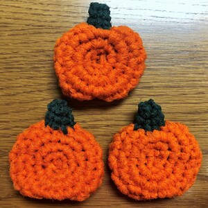 May include: Three crocheted orange pumpkins with green stems. The pumpkins are made with a textured yarn and have a circular design.