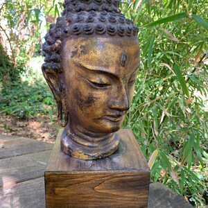 May include: A golden Buddha head sculpture with a dark brown wooden base. The sculpture is detailed with intricate carvings and a serene expression.