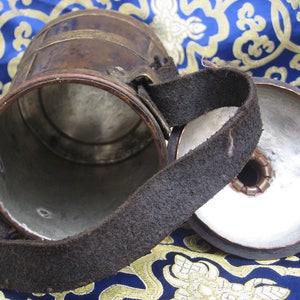 May include: A vintage copper and silver drinking cup with a leather strap handle. The cup has a round, open top and a closed bottom. The cup is sitting on a blue and gold patterned fabric.