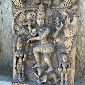 May include: A carved wooden wall hanging depicting a Hindu deity with multiple arms, standing on a smaller figure. The deity is surrounded by intricate floral designs and other figures.