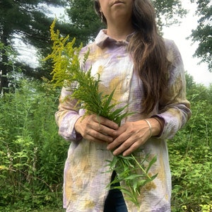 May include: A person wearing a purple and yellow tie-dye shirt with long sleeves and a button-down collar. They are holding a bouquet of yellow wildflowers.