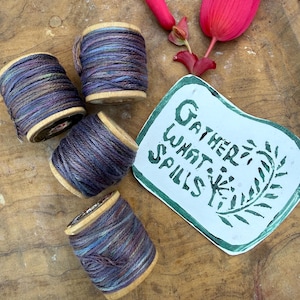 May include: Four wooden spools wrapped in variegated purple, blue, and grey thread, accompanied by a bright pink flower bud and a white paper cutout featuring the green hand-lettered text "GATHER WHAT SPILLS" alongside a botanical illustration.
