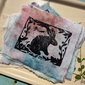 May include: A square fabric piece with a black rabbit print inside a rectangular frame, surrounded by leaves. The fabric has a frayed edge and is dyed with pink, blue, and white hues. The item is likely handmade.