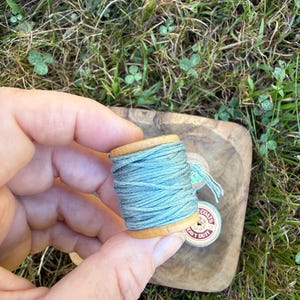 May include: A wooden spool wrapped with light blue thread. The spool has a cylindrical shape with flat ends. A small circular label on the side of the spool features the text COATS and HEAVY DUTY in red and black ink on a white background.