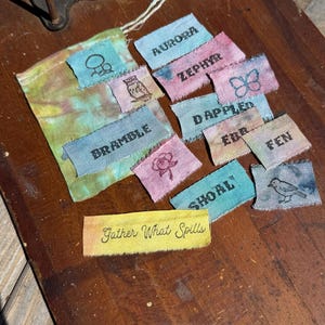 Puede incluir: Una colección de pequeñas etiquetas rectangulares de tela con palabras e imágenes bordadas, incluyendo "Aurora", "Zephyr" y "Gather What Spills". Las etiquetas son de varios colores, como azul, rosa y amarillo, y están sobre una superficie de madera.
