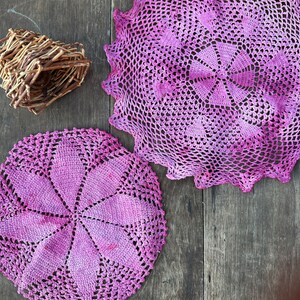 May include: Two pink crocheted doilies on a wooden surface. The doilies have a delicate, lacy design and are made with a soft, yarn.