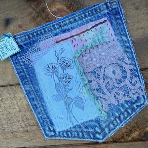 May include: A blue denim pocket featuring a rectangular fabric patch with a floral embroidery design. The pocket is decorated with layered pink lace, a green ruffled fabric strip, and a small tag with the text Gather What Spills.