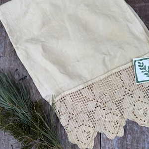 May include: A folded, cream-colored linen textile with a crocheted edge. The crocheted border is a light beige color and has a scalloped design. A sprig of green pine needles is on the left side of the textile.