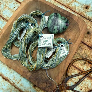 May include: Three skeins of variegated green, blue, and yellow embroidery thread with white paper tags reading Gather What Spills, accompanied by a polished heart-shaped green and black stone, all resting inside a square wooden bowl.