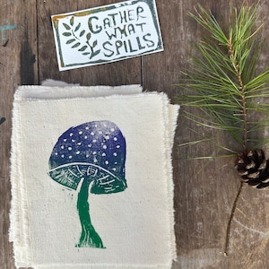 以下が含まれることがあります： 緑と紫のキノコ柄の白い布の正方形が積み重ねられています。布地はほつれた縁を持っています。別々のカードに「Gather What Spills」というテキストが印刷されています。