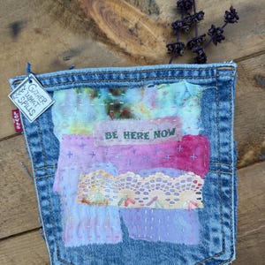 May include: A blue denim pocket featuring a patchwork design of multi-colored fabric scraps, lace trim, and hand-stitched embroidery. The text BE HERE NOW is stitched in green thread. A small tag attached to the side reads GATHER WHAT SPILLS.