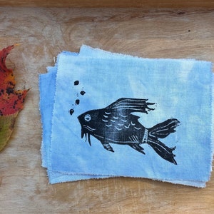 May include: A black and white hand-printed design of a fish on a stack of light blue fabric squares.