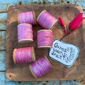 May include: Five wooden spools wrapped in variegated pink thread, a single bright pink tulip flower, and a white rectangular tag with rounded corners featuring the hand-lettered black text "Gather What Spills" and a small botanical illustration.