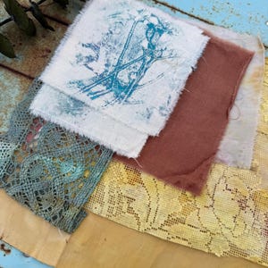 May include: Assortment of fabric swatches in various colors and textures. Includes a white square with a blue botanical print, a rust-colored piece, and sections of lace in teal and yellow. The fabrics are arranged on a weathered, blue-painted surface.