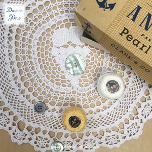 May include: A white crochet doily with a yellow and white spool of thread, a white spool of thread, a blue button, and a mother-of-pearl button. The doily is on a brown surface. The spools of thread have the text "Anchor Pearl Cotton" on them.