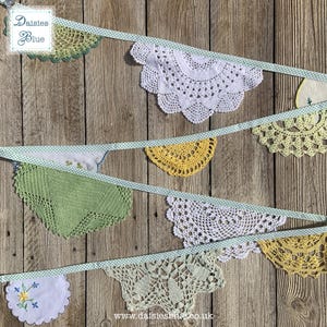 May include: A handmade bunting banner featuring various crochet doilies in white, yellow, and green, strung on a light blue ribbon with white polka dots. The banner is displayed against a rustic wooden background. The banner has the text "Daisies Blue".