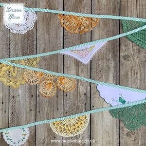 May include: A banner of vintage doilies in shades of white, yellow, and green, strung on a light blue and white striped ribbon. The doilies are of various shapes and sizes, with intricate lace patterns. The background is a rustic wooden surface. The text "Daisies Blue" is in the upper left corner.