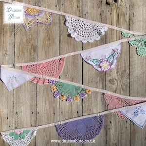 Può includere: Una ghirlanda di bandierine colorata realizzata con centrini e fazzoletti vintage all'uncinetto e ricamati. La ghirlanda presenta vari colori, tra cui rosa, verde, viola e bianco, su uno sfondo di legno rustico. La ghirlanda è infilata su un nastro a righe.