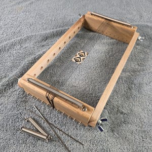May include: A wooden weaving loom with metal springs and needles. The rectangular frame has a natural wood finish. A small woven piece is in the center. Additional metal springs and needles are placed nearby.