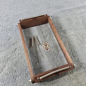 May include: A rectangular wooden bead loom featuring a dark brown wood frame with two horizontal metal springs attached to the top and bottom bars. The loom includes thin vertical warp threads and two metal needles, with a small woven bead pattern.