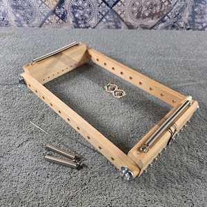May include: A rectangular wooden bead loom featuring a light-colored wood frame with metal springs at both ends. Thin black warp threads are strung across the frame, holding a small partially woven beaded pattern. Two extra metal springs are included.