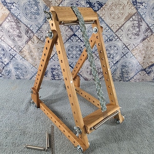 May include: A wooden loom with an A-frame structure featuring adjustable side rails with multiple holes, metal wing nuts, and tension springs. A green and white patterned woven band is attached to the top and bottom bars. Two extra metal springs and a thin metal rod lie separately.