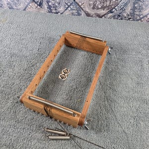 May include: A rectangular wooden bead loom featuring a light brown wood frame, two metal tension springs at the ends, and thin vertical warp threads. A small, partially woven geometric bead pattern in black and white sits in the center, with two additional loose metal springs and thin wires placed below the frame.