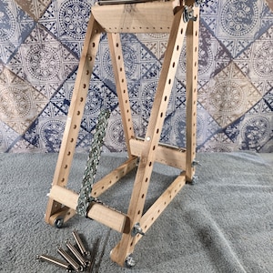 May include: A wooden bead loom with a triangular frame, featuring a partially woven green and white patterned bracelet. The loom includes metal hardware, wing nuts, and a tension spring. Four additional metal springs and two thin needles lie nearby.