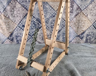 May include: A wooden bead loom with a triangular frame, featuring a partially woven green and white patterned bracelet. The loom includes metal hardware, wing nuts, and a tension spring. Four additional metal springs and two thin needles lie nearby.