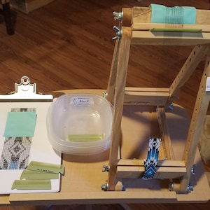 May include: A wooden bead loom with a clipboard, plastic container, and various craft supplies. The clipboard holds a partially beaded piece and a teal square. The loom has a teal thread and a beaded design.