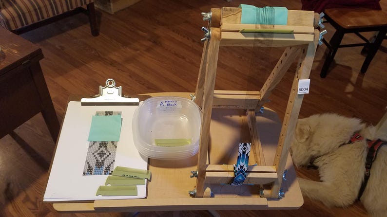 May include: A wooden bead loom with a clipboard, plastic container, and various craft supplies. The clipboard holds a partially beaded piece and a teal square. The loom has a teal thread and a beaded design.
