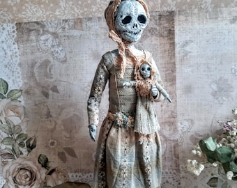 Może przedstawiać: Ręcznie robiona porcelanowa lalka z czaszką i długą, falującą suknią. Lalka trzyma w ramionach mniejszą porcelanową lalkę. Lalka stoi na czarnej podstawie.