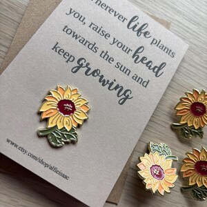 Op de afbeelding: Zonnebloem emaille pins met gele, oranje en rode bloemblaadjes, groene bladeren en een bruin centrum. De pins liggen op een kaart met de tekst "Wherever life plants you, raise your head towards the sun and keep growing."