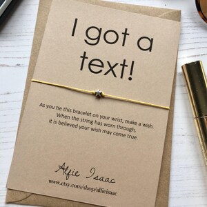 May include: A greeting card with the text "I got a text!" and a gold string bracelet with a star charm. The card is on a brown envelope. The text on the card reads, "As you tie this bracelet on your wrist, make a wish."