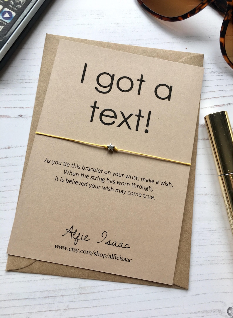 May include: A greeting card with the text "I got a text!" and a gold string bracelet with a star charm. The card is on a brown envelope. The text on the card reads, "As you tie this bracelet on your wrist, make a wish."