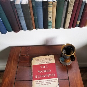Puede incluir: Una colección de libros antiguos en una estantería sobre una mesa de madera. Sobre la mesa, un libro titulado "The World Remapped" y una taza de té. El libro tiene una cubierta roja con texto blanco. Los libros son de varios colores.