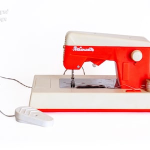 Op de afbeelding: Een vintage MaCousette naaimachine met een crèmekleurige en feloranje kunststof behuizing, een metalen steekplaat, een wit voetpedaal verbonden met een snoer en een kleine houten garenklos aan de rechterkant van de machinebasis.