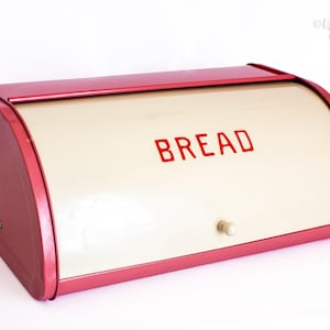 Może przedstawiać: Prostokątny metalowy chlebak z czerwoną ramą i kremowymi drzwiczkami przesuwnymi. Słowo BREAD jest nadrukowane czerwonymi wielkimi literami na przednim panelu. Małe okrągłe kremowe pokrętło jest przymocowane do środka drzwiczek.