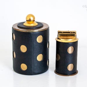 Pode incluir: Um conjunto vintage de recipiente e isqueiro em couro sintético preto. As peças cilíndricas apresentam detalhes dourados e recortes circulares. O recipiente tem uma tampa e um botão dourados. O isqueiro tem um topo dourado.