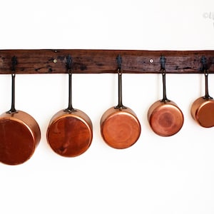 May include: A set of five graduated copper saucepans with long metal handles hanging from a dark, rough-hewn wooden wall rack. The pans are arranged in descending order of size from left to right, featuring polished metallic surfaces.