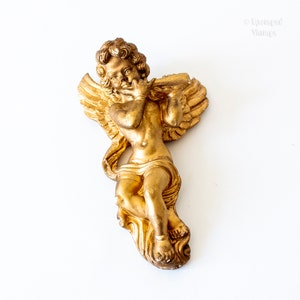 Può includere: Una statuetta di cherubino dorata che suona il flauto. Il cherubino ha le ali ed è raffigurato in una posa di volo. La statuetta è realizzata in un materiale liscio e lucido.